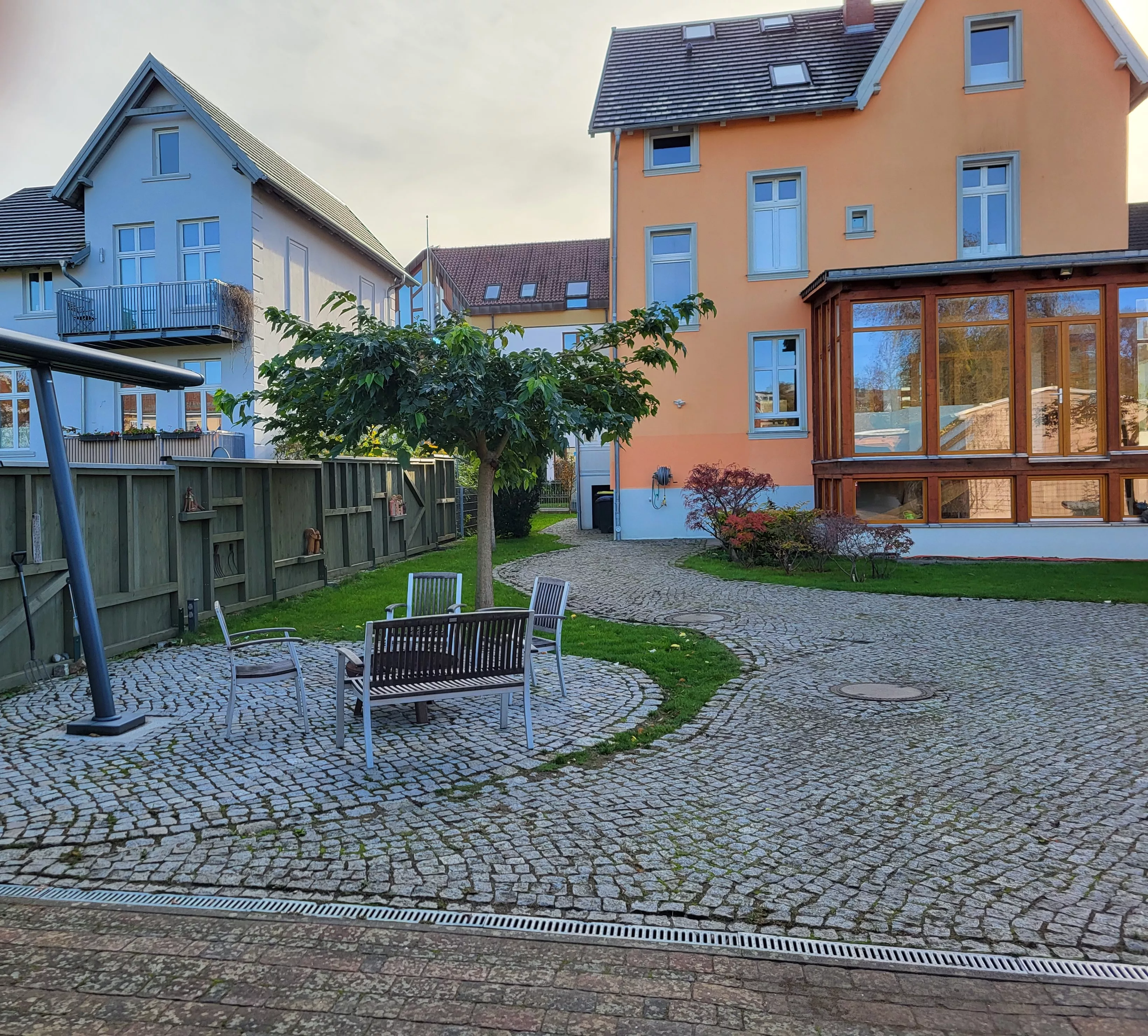 Gepflasterter Innenhof mit Holzbank, Stühlen aus Metall und Baum sowie Blick auf Nachbarhäuser mit Wintergarten