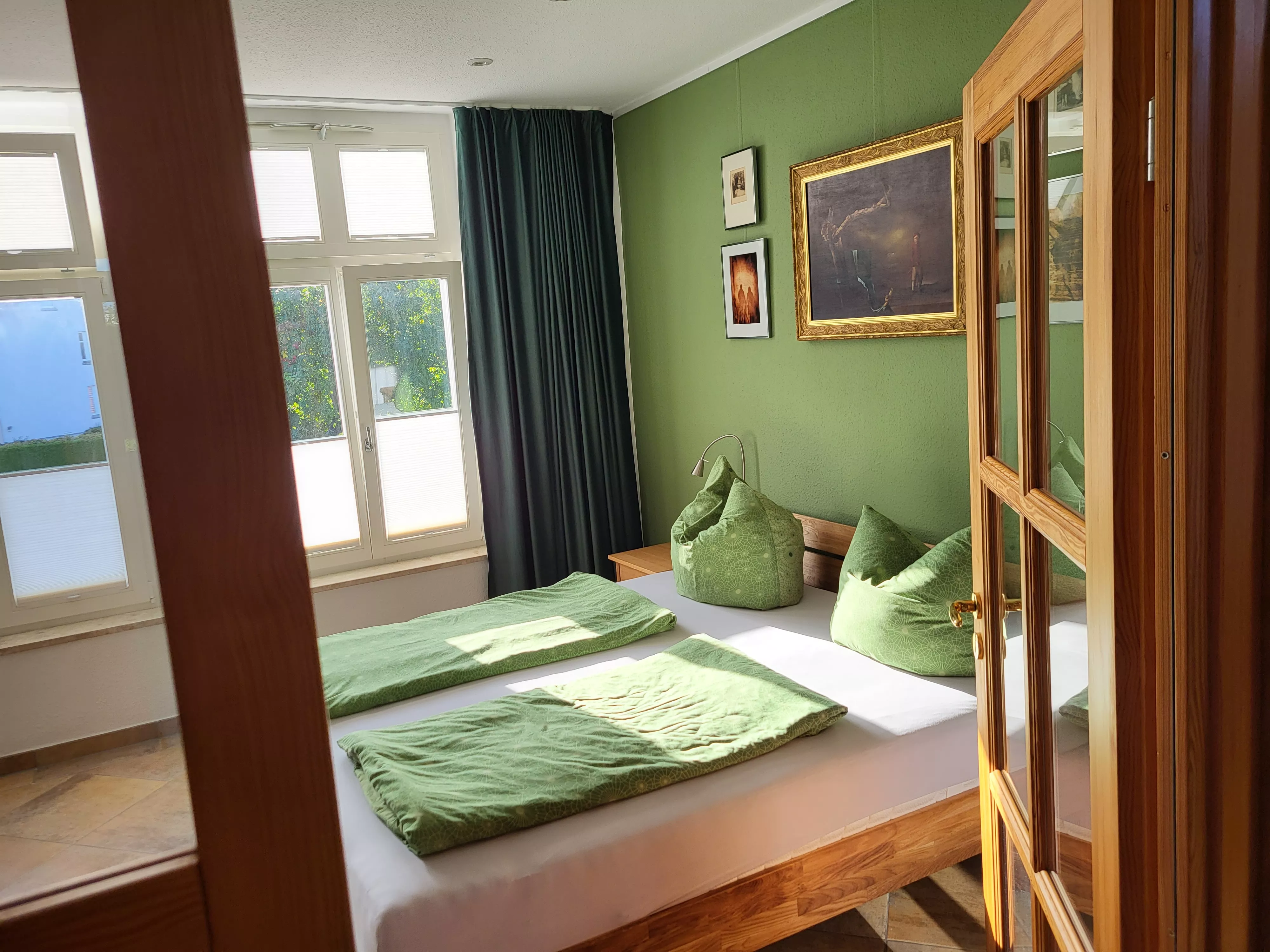 Schlafzimmer mit Doppelbett aus Holz, grüner Bettwäsche, grüner Wand mit gerahmten Kunstdrucken und Sprossentür aus Holz